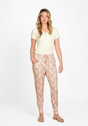 Vrouw staand, gekleed in een lichtbeige top met korte mouwen, broek met bloemmotief en trekkoord, en beige open teen slippers op een witte achtergrond.