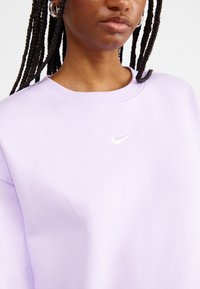 Sweatshirt lilás claro com gola redonda, punhos canelados e um pequeno logotipo branco da Nike no peito. Tecido suave.