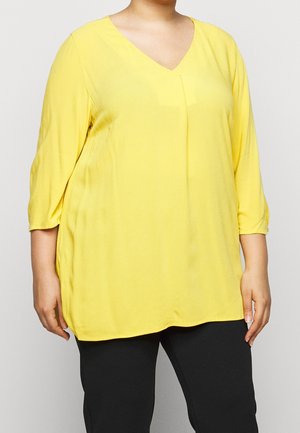 Persona con una blusa amarilla holgada con cuello en V y mangas tres cuartos, y pantalones negros, de pie sobre un fondo liso.