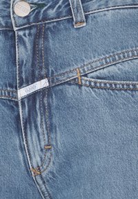 Gros plan sur un jean en denim bleu montrant la ceinture, la passants de ceinture, la poche avant et un bouton en métal portant l'inscription « CLOSED ».