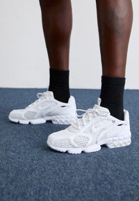 Bílé sportovní boty vyrobené z materiálu síťoviny a syntetiky, s texturovaným designem, šněrováním vpředu a výraznou vzduchovou podrážkou.