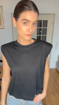 Νεαρή γυναίκα με καστανά μαλλιά πιασμένα πίσω, φορά μαύρο αμάνικο τοπ και ανοιχτό μπλε τζιν, στέκεται σε φωτεινό δωμάτιο με ξύλινο πάτωμα.