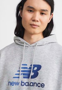 Siva kapuca majica iz bombaža s teksturiranim finišem. Na prednji strani se nahaja modri logotip "New Balance". Kapuca ima prilagodljive vrvice.