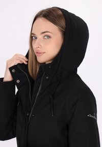 Zwarte hoodie met ritssluiting en drukknoopsluiting, met een textuur buitenkant, verstelbare trekkoord en logo detail op de mouw.