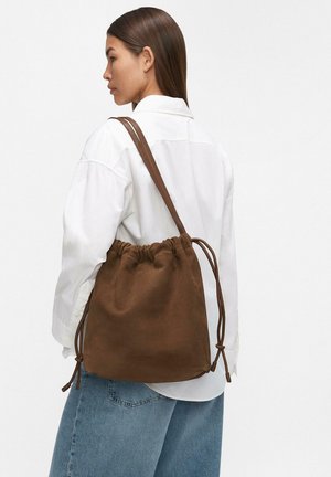 Borsa a tracolla in suede marrone con lacci accentuati, texture morbida e forma sciatta, indossata sopra una camicia bianca e jeans blu.