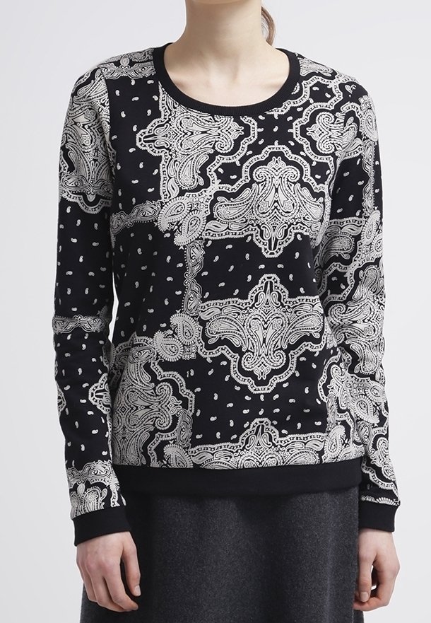 Haut noir à manches longues avec des motifs paisley blancs complexes et des motifs en pointillé, porté par une personne ayant les mains détendues le long du corps.