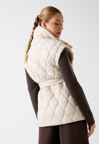 Gesteppte cremefarbene Weste mit hohem Kragen, tailliertem Gürtel und diamantförmiger Struktur, getragen über einem braunen Oberteil. Rückansicht gezeigt.