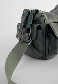Sac à épaule noir avec une large sangle grise réglable et une boucle métallique, avec des poches latérales et des coutures visibles sur un matériau lisse.
