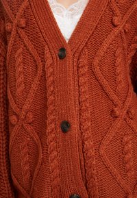 Rdzawoczerwony sweter z dzianiny z głębokim dekoltem w kształcie litery V, teksturowanymi warkoczami i dużymi czarnymi guzikami. Teksturowany wzór zawiera wypukłe wzory.