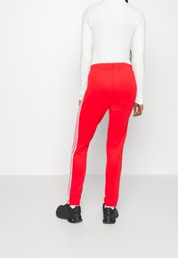 Mujer vestida con pantalones deportivos rojos con franjas blancas a los lados, camiseta de manga larga blanca y zapatillas deportivas negras, de pie contra un fondo blanco liso.