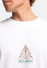 White cotton T-shirt featuring a geometric brown and blue graphic design on the chest, with a triangular shape and abstract patterns.