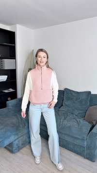 Rózsaszín és krémszínű zippzáras pulóver, puha anyagból készült, világos kék, bő szárú farmernadrággal párosítva; magas gallérral és bordázott mandzsettákkal rendelkezik.