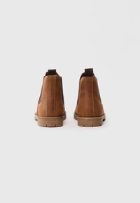 Une paire de bottes en cuir marron à bout arrondi avec des panneaux latéraux en élastique noir, vues de derrière sur un fond blanc.