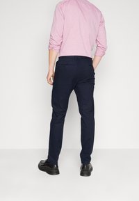 Pantaloni sartoriali blu navy con una texture liscia, abbinati a una camicia a righe rosa e scarpe eleganti nere. Vita e tasche visibili.