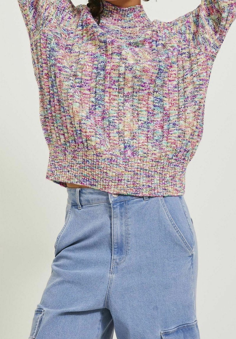 Femme portant un pull en maille multicolore avec les bras levés et un pantalon en jean bleu clair taille haute avec des poches avant.