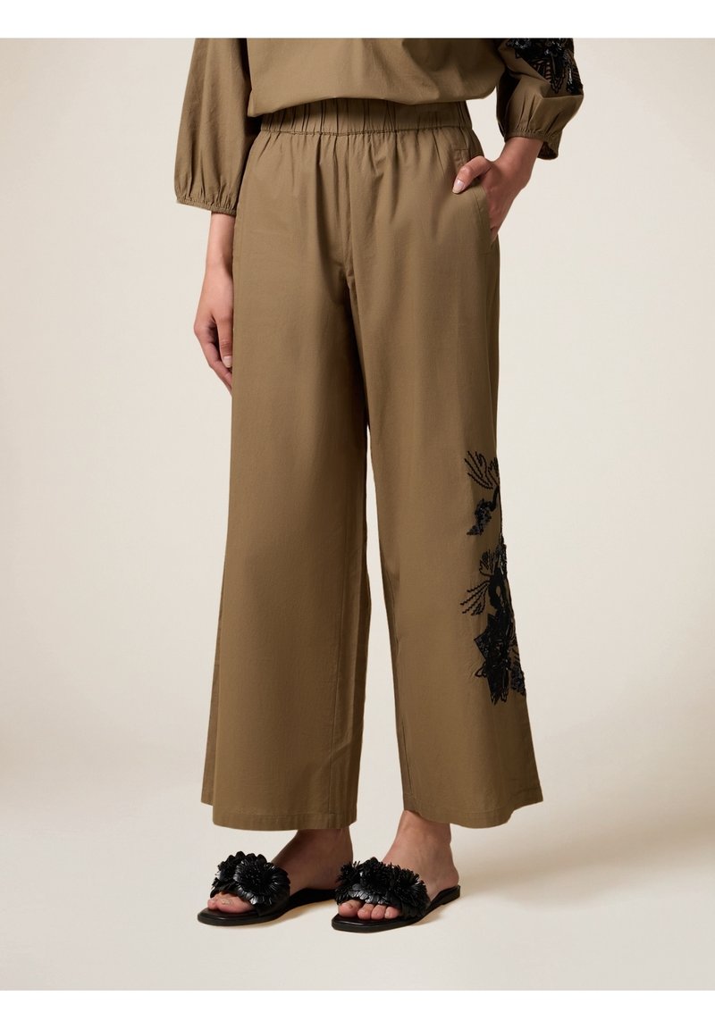 Lichte, olijfgroene, wijde broek met geborduurd zwart bloemenpatroon aan de linkerkant, voorzien van een elastische tailleband en zijzakken.