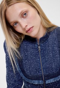 Temno modra denim jakna z zadrgo spredaj, zaključki z resicami in teksturirano, pikčasto tkanino. Visok ovratnik in oblikovan dizajn.