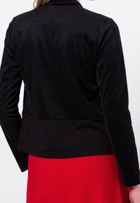 Veste en daim noir avec un design structuré et une texture douce, dotée d'un col haut et d'une taille cintrée, associée à un vêtement rouge en dessous.