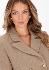 Cappotto beige su misura con design doppiopetto, colletto a punta e accenti in bottoni argentati lucidi, realizzato in un misto di lana testurizzata.