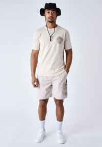 Camiseta crema con un logo, pantalones cortos beige con un diseño estampado, zapatillas blancas y un sombrero tipo bucket negro. Atuendo simple y casual.