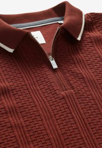Polo en maille couleur rouille avec motifs verticaux texturés, col bordé de blanc et fermeture éclair métallique partiellement fermée au niveau du cou.