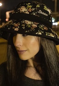 Cappello a secchiello nero con motivo floreale e ampia visiera, caratterizzato da un nastro in velluto e un logo ricamato "Blackskies" sulla parte anteriore. Sfondo scuro con luci urbane.
