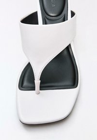 Hvid læder-tåsandal med firkantet tå og sort polstret fodseng, der viser detaljeret syning på remmen.