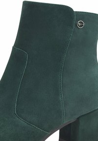 Stivale alla caviglia in suede verde con tacco a blocco, dettagli di pannelli cuciti e un piccolo accento metallico vicino all'apertura.