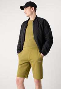Crna bomber jakna, maslinasto zelena uska majica, i odgovarajuće kratke hlače s logom. Jednostavan dizajn, glatka tekstura i ležeran sportski stil.