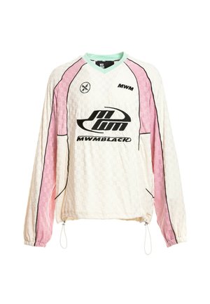 Joven con camiseta de manga larga blanca y rosa, pantalones cortos beige con detalle de cadena, calcetines blancos y zapatos beige texturizados sin cordones, de pie.