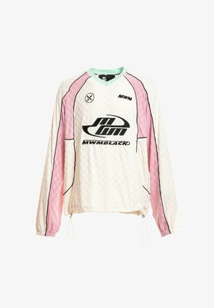 Joven con camiseta de manga larga blanca y rosa, pantalones cortos beige con detalle de cadena, calcetines blancos y zapatos beige texturizados sin cordones, de pie.