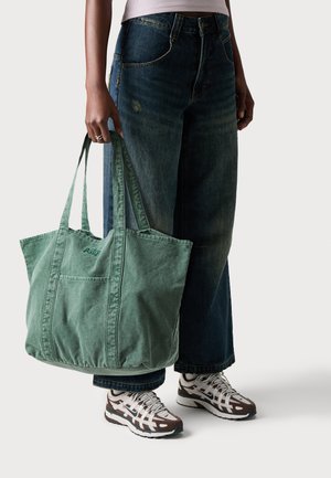 Borsa tote in cotone verde con dettagli cuciti e una tasca frontale, tenuta da una persona che indossa jeans larghi blu scuri e scarpe da ginnastica con motivo.