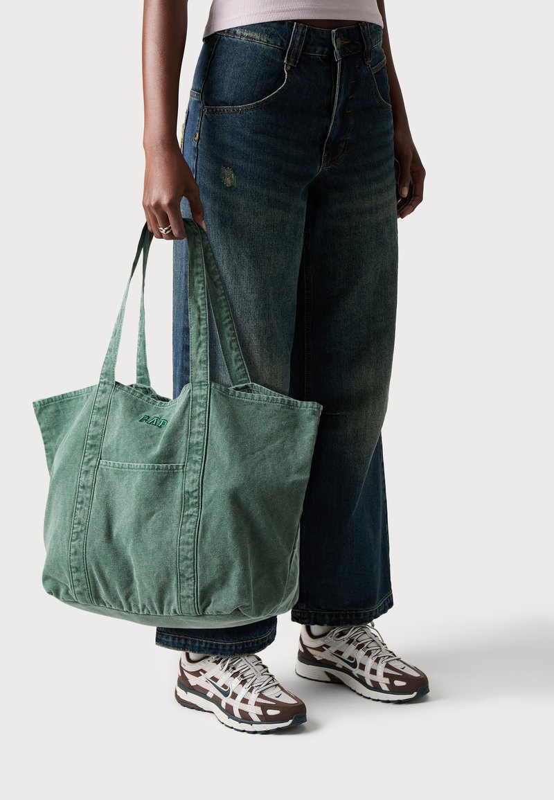 Grüne Baumwolltasche mit gesteppten Details und einer Fronttasche, gehalten von einer Person, die dunkelblaue weit geschnittene Jeans und gemusterte Sneakers trägt.