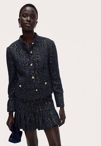 Veste en tweed bleu marine avec boutons dorés et jupe assortie à volants. Le tissu présente un aspect texturé avec des détails scintillants subtils.