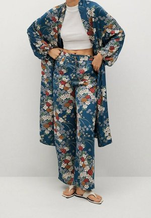 Osoba ubrana w niebieskie kwiatowe kimono i pasujące spodnie, biała krótka bluzka i białe sandały, stojąca na jednolitym tle.