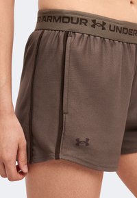 Κοντινή λήψη καφέ σορτς Under Armour με ελαστική μέση και λογότυπο στο κάτω αριστερό μηρό, φορεμένα από ένα άτομο που στέκεται με χαλαρό χέρι.
