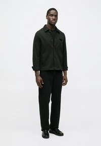 Must musta nööbitav jakk pika varrukaga, kantud musta särgi kohal, ühendatud mustade laiade pükste ja mustade kingadega, kõik minimalistlikus stiilis.