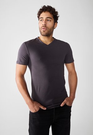 Mørk grå v-hals t-shirt lavet af blødt stof med korte ærmer, parret med sorte jeans, hænderne placeret i lommerne.