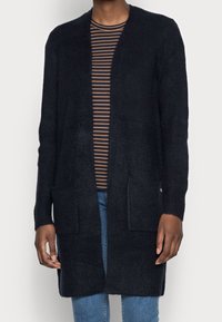 Pitkä, tummansininen neuletakki, joka on valmistettu teksturoidusta neulekankaasta. Ominaisuuksina avonainen etuosa, kaksi etutaskua ja ribbireunat. Päällä raidallinen paita.