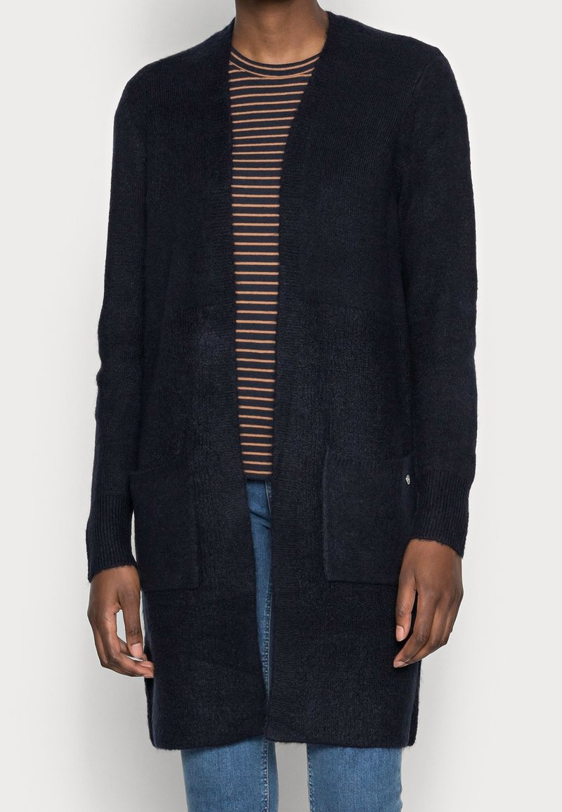 Pitkä, tummansininen neuletakki, joka on valmistettu teksturoidusta neulekankaasta. Ominaisuuksina avonainen etuosa, kaksi etutaskua ja ribbireunat. Päällä raidallinen paita.