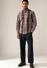Camisa de cuadros marrón con líneas blancas y verde oscuro, combinada con pantalones azul marino y zapatos negros, de pie sobre un fondo claro.