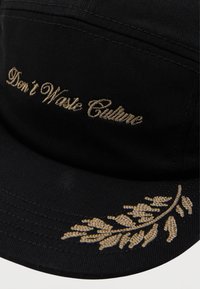 Svart caps laget av stoff, med gullbroderi av teksten "Ikke kast bort kulturen" og et bladdesign. Strukturert form med justerbar stropp.