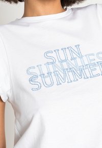 Camiseta de algodón blanca con texto de rhinestones azules que dice "SOL GAFAS VERANO" en diferentes fuentes y tamaños, con mangas cortas.