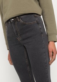 Jeans ajustados negros con una textura suave, con costuras naranjas y diseño clásico de cinco bolsillos. Combinados con una camiseta de manga larga gris.