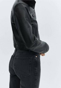 Czarna skórzana kurtka typu cropped z sztucznym futrem przy kołnierzu, zestawiona z czarnymi dżinsami o wysokim stanie. Wyróżnia się gładką fakturą i dopasowanym krojem.