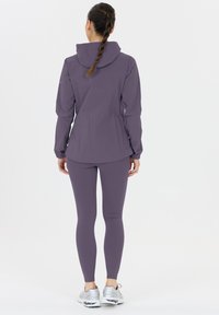 Vrouw met gevlochten haar, gekleed in een paarse gewatteerde jas met capuchon, bijpassende leggings en zilveren sneakers, staand met de rug naar de camera op een witte achtergrond.