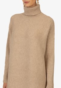Pull à col roulé en tricot beige avec une texture en relief horizontale, coupe décontractée et manches longues, présentant un col haut et un tissu doux.