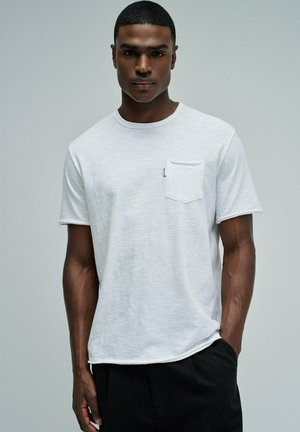 Joven con el pelo corto que lleva una camiseta blanca lisa con un bolsillo en el pecho y pantalones negros, posando frente a un fondo gris claro.
