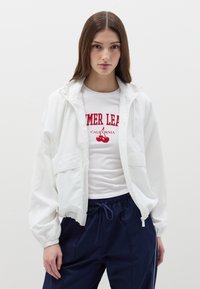 Giacca leggera bianca con polsini elasticizzati e cappuccio, indossata sopra una camicia bianca aderente con testo grafico rosso, abbinata a pantaloni navy.