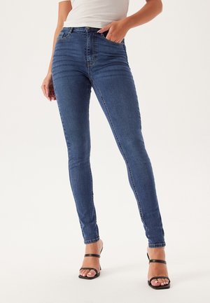 Jeans skinny in denim blu medio, caratterizzati da una vita alta, una silhouette a gamba snella e una leggera scoloritura. Abbinati a tacchi neri con cinturini.